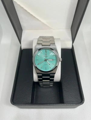 Poedagar silverklocka med turkos urtavla - Snygg klocka från Poedagar med stilren silverlänk och en turkos urtavla med diskret rutmönster. Urtavlan har dag- och datumvisning, och boetten är rund med en modern känsla. Perfekt accessoar för dig som gillar att sticka ut med färg och detaljer.