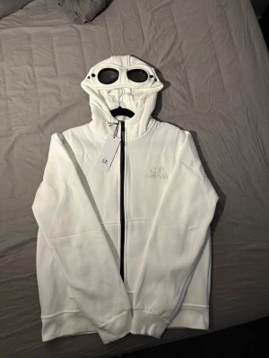Vit C.P. Company Goggle hoodie M - Helt ny storlek M men sitter som S , därav att jag säljer då den va för liten.                                          Vit hoodiejacka från C.P. Company med ikoniska goggle-detaljer i huvan. Jackan har svart dragkedja, broderad logga på bröstet och stora fickor framtill. Tillverkad i mjuk bomull, perfekt för dig som vill sticka ut med streetwear-vibe. 
