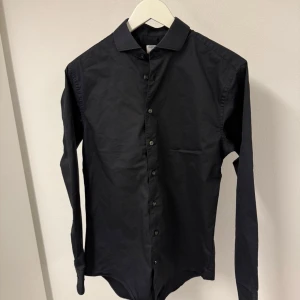 Svart skjorta från Tiger of Sweden - Stilren svart skjorta från Tiger of Sweden i slim fit-modell. Skjortan har klassisk krage, långa ärmar och knappar hela vägen framtill. Materialet är mjuk bomull som känns skönt mot huden. Perfekt för dig som gillar en clean och modern look. 