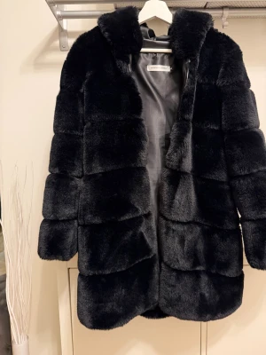 Svart pälsjacka från Borninstockholm - Säljer en svart pälsjacka från Borninstockholm med fluffig känsla och huva. Jackan har ett stilrent, blockindelat mönster och är fodrad med slätt tyg på insidan. Perfekt för dig som vill ha en varm och snygg look under kyliga dagar. Liten i storleken! 