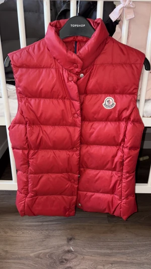 Röd dunväst från Moncler - Riktigt snygg röd dunväst från Moncler med hög krage och klassisk logga på bröstet. Västen är quiltad och har tryckknappar framtill. Perfekt för lager-på-lager och ger en cool vibe till din outfit.