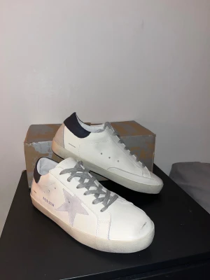 Golden Goose vita sneakers med grå stjärna - Golden Goose sneakers i vitt skinn med grå mockadetaljer och ikonisk stjärna på sidan. Grå snörning,  mörkblått hälparti och platt sula. Snygg och stilren modell som passar perfekt till jeans eller shorts. Klassisk low-top design med coola detaljer.