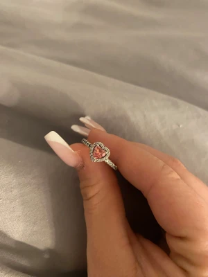Silverring med rosa hjärtformad sten från pandora - Superfin silverring med en gnistrande rosa hjärtformad sten i mitten, omgiven av små klara stenar runt hela hjärtat och längs ringen. Perfekt för dig som gillar söta detaljer och vill ha något som sticker ut lite extra.