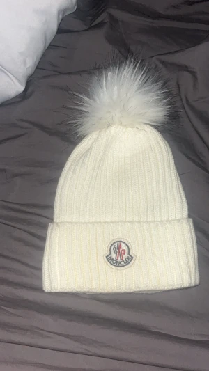 Vit Moncler mössa med pälsboll - Snygg vit ribbstickad mössa från Moncler med stor fluffig pälsboll på toppen och klassisk Moncler-logga framtill. Perfekt för kalla dagar när du vill ha både stil och värme. Mössan har uppvikt kant och känns riktigt lyxig.