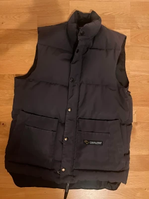 Svart dunväst från Canada Goose XS - Svart dunväst från Canada Goose i storlek XS. Västen har hög krage, två stora fickor framtill med tryckknappar och logotyp på ena fickan. Quiltad design och dragkedja med knappslå. Perfekt för kalla dagar när du vill hålla värmen och ändå se chill ut. Den är ett gammalt modell men i en jättebra kondition och inga skador.
