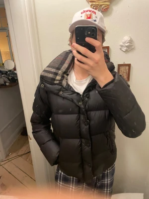 Svart Burberry dunjacka med rutig huva - Säljer en svart pufferjacka från Burberry med ikonisk rutig huva. Jackan har dragkedja och knappar framtill, samt två sidofickor med dragkedja. Materialet är polyamid och fyllningen består av grågåsdun. Perfekt för kalla dagar och har en clean, klassisk look.