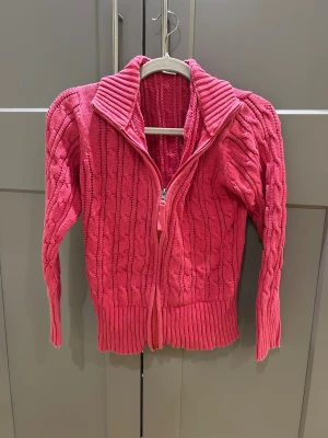 Rosa kabelstickad kofta från Isolde - Snygg rosa kabelstickad kofta från Isolde med dragkedja framtill och bred ribbad krage. Tröjan har långärmad design och ribbade muddar vid ärmslut och nederkant. Perfekt för dig som gillar färg och vill ha en mysig look.