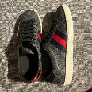Gucci ace - Snygga Gucci sneakers i mörkgrått med GG-monogram över hela skon. Sidan har klassiska ränder i rött och mörkblått, alla tillbehör följer med