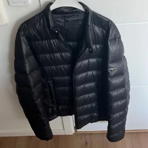 Svart dunjacka från Prada - Snygg svart dunjacka från Prada med klassisk quiltad design och ikonisk logga på ärmen. Jackan har dragkedja framtill, hög krage och praktiska fickor med tryckknapp. Perfekt nu till vintern för dig som vill ha en stilren och lyxig look. Kom gärna med bud. 