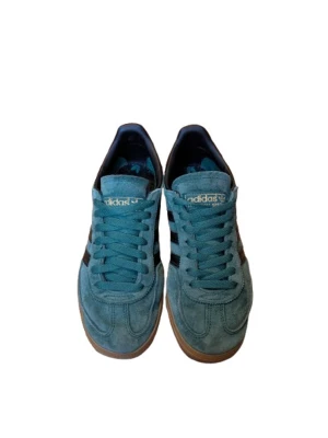 Adidas Spezial gröna sneakers mocka - Adidas Spezial sneakers i grön mocka med svarta läderdetaljer och klassiska tre ränder på sidan. Skorna har snörning, gummisula i beige och guldfärgad Adidas-logga på häl och plös. Rund tå och låg profil ger en clean och retro vibe. Saknar låda.