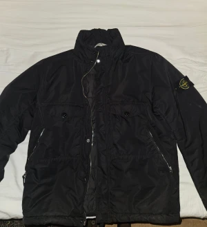 Svart dunjacka från Stone Island - Svart dunjacka från Stone Island med ikonisk patch på vänster ärm. Jackan har hög krage, två bröstfickor med knappar och två sidofickor med dragkedja. Insidan är quiltad och jackan stängs med både dragkedja och knappar. Perfekt för kalla dagar. 
