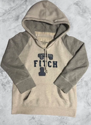 Grå hoodie Abercrombie & Fitch L - Snygg hoodie från Abercrombie & Fitch i storlek L i Barn Tröjan är ljusgrå med mörkare grå ärmar och har ett stort tryck med texten 'FITCH' och broderade detaljer i blått på bröstet. Utrustad med huva och snörning samt en klassisk magficka framtill.Passar XS i herr storlek.