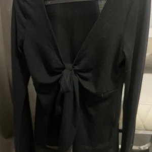 Svart omlottopp med knyt framtill - Svart långärmad omlottopp med djup v-ringning och knyt framtill. Toppen är croppad och har en tight passform som framhäver midjan. Perfekt för dig som gillar en stilren och trendig look. Suddiga bilder då min kamera är sönder 😊