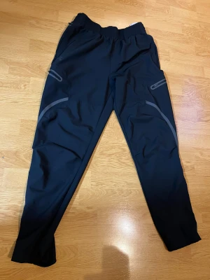 Svarta träningsbyxor från Under Armour - Snygga svarta träningsbyxor från Under Armour med elastisk midja och grå detaljer längs benen. Byxorna har dragkedjeförsedda fickor på sidorna och är tillverkade i ett lätt syntetmaterial som passar perfekt för aktiva dagar.