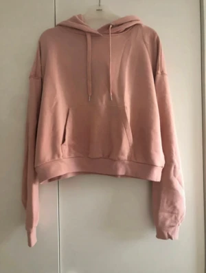 Croppad puderrosa hoodie med huva - Säljer en croppad hoodie i puderrosa med huva och snörning. Tröjan har en stor magficka och långa ärmar med ribbade muddar. Tröjan har en avslappnad passform Perfekt och är  enkel att matcha med jeans eller leggings