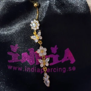 Lång navelpiercing guld - Fin lång navelpiercing i kirurgiskt stål, guldpläterat. Passar dig som kanske är känslig mot annan metall. Aldrig använd. Fjärilar och blommor på hänget. 
