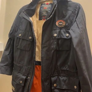 Morris marinblå parkajacka med huva - Parkajacka från Morris i marinblått med huva och flera praktiska fickor framtill. Jackan har snygga detaljer som patch med '1968' på bröstet, dragkedja och tryckknappar. Perfekt för dig som gillar sportig och klassisk stil. Jackan har storlek S men sitter som M