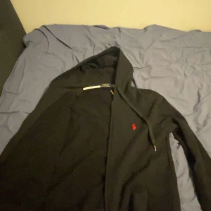 Svart hoodie från Ralph Lauren - Svart hoodie från Ralph Lauren med dragkedja, vit snörning och röd broderad logga på bröstet. Klassisk passform, huva och kängurufickor framtill. Perfekt för en chill och stilren look.