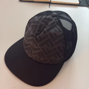 Svart Fendi keps med mesh och logga - Snygg svart keps från Fendi med mesh på sidorna och bak, samt ett geometriskt mönster framtill. Justerbar med guldfärgad Fendi-logga bak. Tillverkad i 100% polyester, perfekt för dig som gillar exklusiva accessoarer med streetkänsla.
