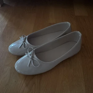 Beige ballerinaskor med rosett från Nelly - Snygga beige ballerinaskor från Nelly med glansig finish och rund tå. Skorna har en söt rosett framtill och platt sula, vilket gör dem både stilrena och bekväma. Perfekta för dig som gillar enkel och chic stil. Helt oanvända.