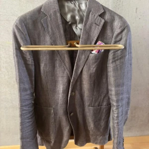 Grå brun linnékostym Oscar Jacobson - Snygg tvådelad kostym från Oscar Jacobson i 100% linne från Angelico. Kostymen är brun med klassisk kavaj med två knappar, utanpåfickor och matchande byxor. Perfekt för dig som vill ha en stilren och luftig look.