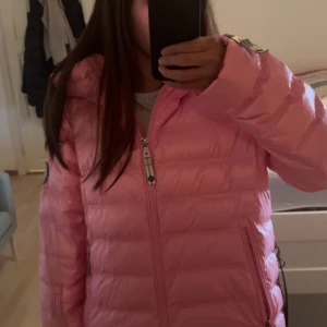 Rosa pufferjacka från CAYA - Säljer en riktigt snygg rosa pufferjacka från CAYA. Jackan har glansig finish, huva, dragkedjor med coola detaljer och ett stort logomärke på insidan. Perfekt för dig som vill sticka ut med färg i vinter eller höst. Helt ny! 