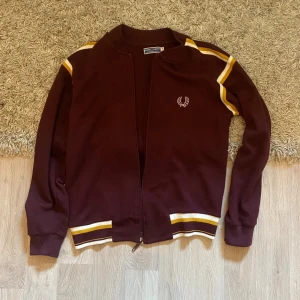 Vinröd Fred Perry kofta med gula ränder - Säljer en vinröd kofta från Fred Perry med dragkedja och vita samt gula ränder på axlar och muddar. Klassisk logga broderad på bröstet. Perfekt för dig som gillar sportig och retro stil.