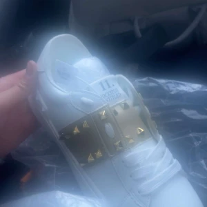 Valentino Rockstud sneakers vit/guld - Snygga Valentino Rockstud sneakers i vitt skinn med guldiga detaljer och ikoniska nitar över snörningen. Platt sula med mönstrad kant och rund tå. Märket syns tydligt på plösen och skon har en lyxig vibe med metallic och nitar.