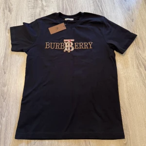 Svart Burberry t-shirt M - Svart t-shirt från Burberry med broderad logga i guld och beige på bröstet. Klassisk rund hals och korta ärmar. Tillverkad i mjuk bomull med lite stretch för extra komfort. Snygg detalj med det ikoniska Burberry-mönstret i bokstaven B.