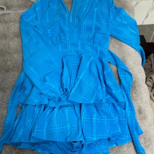 Blå rutig kortklänning från Amisu - Supersöt blå kortklänning från Amisu med rutigt mönster, långa ärmar och v-ringning. Klänningen har volangdetaljer och ett brett knytband i midjan som ger en snygg siluett. Perfekt till festen evenemang eller middagar❤️