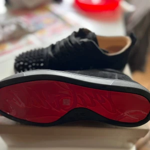 Christian Louboutin svarta sneakers med nitar - Svarta sneakers från Christian Louboutin med klassisk röd sula och svarta nitar på tån. Skorna har snörning och är tillverkade i mocka med läderdetaljer. Snygg och edgy design som sticker ut med den ikoniska röda sulan och coola detaljer.