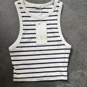 Randig croppad linnetopp från Zara - Säljer en fräsch croppad linnetopp från Zara i vitt med svarta horisontella ränder. Toppen är ribbad, ärmlös och har en tight passform med rund halsringning. Oanvänd pga jag inte gillar modellen.