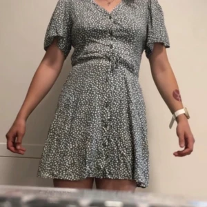 Blommig kortklänning från Vero Moda - Supersöt kortklänning från Vero Moda med blommigt mönster i vitt på gråblå botten. Klänningen har v-ringning, knappar framtill och korta puffärmar. Perfekt för sommarens alla tillfällen och har en lätt, flowy passform.