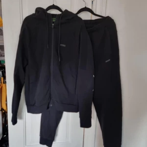 Svart dress från BOSS - Svart dress från BOSS med dragkedja framtill och huva med snörning. Tröjan har en clean look med diskret BOSS-logga på bröstet och ribbade muddar. Byxorna dess lika. Perfekt för chill dagar eller när du vill ha en stilren streetwear vibe. 