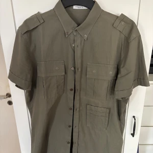 Olivgrön skjorta från J.Lindeberg (Strl 48/S) - Snygg olivgrön skjorta från J.Lindeberg med korta ärmar, axelklaffar och tre fickor framtill. Skjortan har klassisk krage och knäppning med knappar hela vägen. Perfekt för dig som gillar utility-vibbar och stilren design.