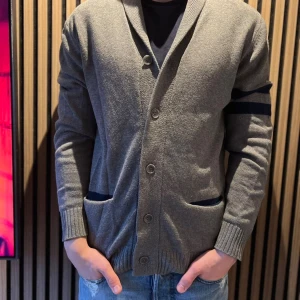 Grå kofta från Bondelid med blå detaljer - Snygg grå kofta från Bondelid i storlek M. Koftan har två mörkblå ränder på ena ärmen, ribbade muddar och knappar framtill. Fickor med blå insida och en bred krage ger en chill och stilren vibe. Perfekt för lager-på-lager under höst och vinter.