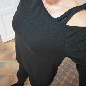 Svart Långärmad Tröja - Cold Shoulder / One Shoulder Cutout M - Säljer en stilren och svart långärmad topp med en iögonfallande axeldetalj.  Den här tröjan har en elegant båthals (bateau neck) och en trendig cut-out över axeln, där en extra tygrem ger en asymmetrisk och intressant detalj. Perfekt för en utekväll eller till jobbet under en kavaj.