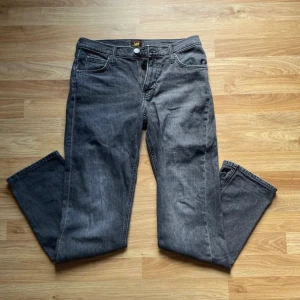 Mörkgrå jeans från Lee - Säljer ett par mörkgrå jeans från Lee med klassisk femficksdesign och raka ben. Jeansen har Lee-logga i midjan och är tillverkade i slitstarkt denimtyg. Perfekta för dig som gillar en enkel och tidlös look. L32