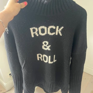 Svart polotröja med Rock & Roll-tryck - Svart stickad polotröja med vit text 'ROCK & ROLL' framtill. Tröjan har en hög, ribbad krage och långa ärmar med breda muddar. Oversized passform som ger en chill vibe, perfekt för dig som gillar statement-plagg och vill sticka ut.