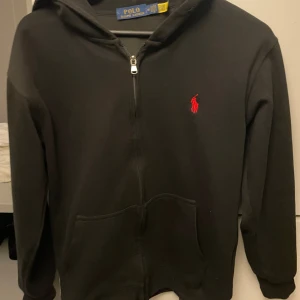 Svart hoodie från Polo Ralph Lauren - Svart hoodie från Polo Ralph Lauren med dragkedja och huva. Klassisk röd logga broderad på bröstet och två stora fickor framtill. Skön passform och perfekt för chill dagar. Materialet är mjuk bomull. Det står M men passar S 