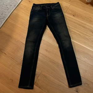 Mörkblå jeans från Nudie Jeans - Snygga mörkblå jeans från Nudie Jeans med klassiska orange sömmar och diskret slitning på benen. Jeansen har fem fickor och är tillverkade i stretchig bomull för en bekväm passform. 