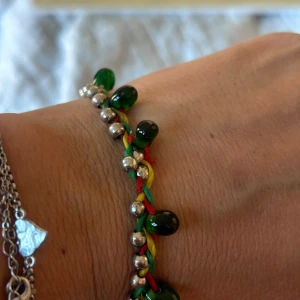 Färgglatt armband med gröna pärlor - Unikt armband med flätad tråd i rött, gult och grönt, dekorerat med gröna glaspärlor och små silvriga kulor. Stängs med en knapp i mörkare ton. Perfekt för dig som gillar färg och boho-stil.