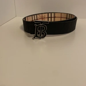 Burberry rutigt skärp med B-spänne - Snyggt Burberry-bälte med klassiskt rutigt mönster i beige, brunt och svart. Bältet har ett stort silvrigt B-spänne framtill och är tillverkat i skinn. Perfekt accessoar för att lyfta din outfit med en lyxig touch går att byta till svart eller beige 