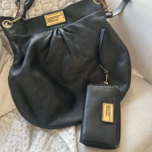 Svart axelväska och plånbok Marc Jacobs - Säljer en snygg svart axelväska från Marc Jacobs i mjukt skinn med guldfärgade metalldetaljer och matchande svart plånbok. Båda har den klassiska Marc Jacobs-plattan framtill. Perfekt storlek för allt du behöver och stilren design. Har ett litet  hål i innerfickan i fodret
