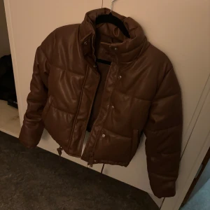 Brun pufferjacka i skinnimitation - Snygg brun pufferjacka med hög krage och dragkedja framtill. Jackan är quiltad och har en cool, boxig passform med dragsko nertill. Tillverkad i skinnimitation som ger en trendig look. Perfekt för dig som vill sticka ut i höst och vinter.