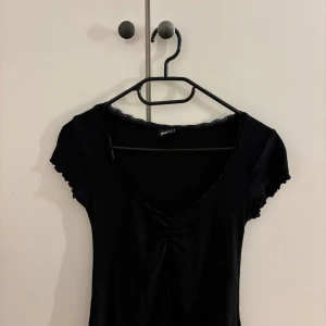 Svart v-ringad topp från Gina Tricot - Svart topp från Gina Tricot med v-ringning och rynkdetalj framtill. Korta ärmar med vågig kant och spetsdetalj längs halsringningen. Mjuk och stretchig bomullskänsla, perfekt till jeans eller kjol.