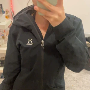 Svart vindjacka från Haglöfs med huva - Säljer en svart vindjacka från Haglöfs i dammodell, storlek S. Jackan har huva, hel dragkedja framtill och två stora fickor. Perfekt för blåsiga dagar och outdoor-aktiviteter. Klassisk och stilren design med Haglöfs-logga på bröstet. Köpt förra året men bara haft på mig den 5 gånger då den var för stor för mig! ( kan gå ner i pris via snabb köp)