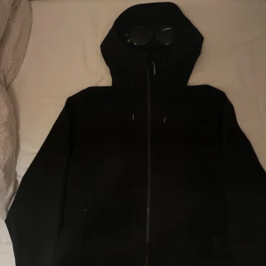 Svart Goggle Jacket från C.P. Company - Svart Goggle Jacket från C.P. Company med ikoniska glasögon i huvan. Jackan har hel dragkedja framtill, två sidofickor med dragkedja och justerbar huva med snörning. Snygg och stilren design, perfekt för dig som gillar streetwear och unika detaljer. Säljer då den ej passar mig oanvänd —- storlek L men mer som S-M