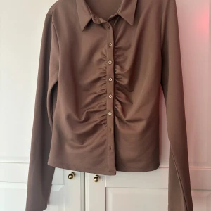 Brun skjorta med rynkade detaljer - Snygg brun skjorta med lång ärm och krage. Framsidan har rynkade detaljer längs knappraden som ger en cool och modern look. Skjortan är tillverkad i ett mjukt syntetmaterial och har en normal passform. Perfekt för dig som gillar stilrena plagg med en twist.