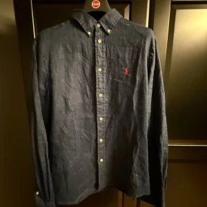 Blå linneskjorta från Polo Ralph Lauren - Snygg mörkblå skjorta från Polo Ralph Lauren i linne med klassisk krage och knappar framtill. Är i storlek XL barnstorlek 18-20 passar någon som är runt 180 och väger mellan 65-80 kg